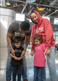 Thumbnail for medium-skinned person wearing a grey crewneck t-shirt with best day of my life written looking down at young child wearing a black crewneck with best day of my life. next to them is a light-skinned person wearing a red pullover hoodie with best day of my life standing behind a young child wearing a pink crewneck t-shirt with best day of my life written on it looking at the floor 