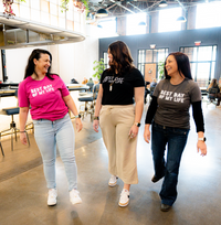 Thumbnail for three light-skinned people with medium to long brown hair looking at each other and laughing. on the left the person is wearing a pink crewneck t-shirt with best day of my life written across in white block lettering, in the center the person is wearing a black v-neck with best day of my life written across in white, on the right the person is wearing a grey crewneck t-shirt with best day of my life written in block lettering over a long-sleeved black shirt.