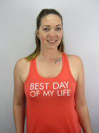 Thumbnail for light-skinned person with shoulder length blonde hair wearing an orange tank top with best day of my life written across in white.