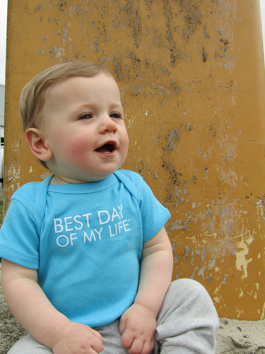 light-skinned infant with short blond hair wearing a sky blue onesie that reads best day of my life 